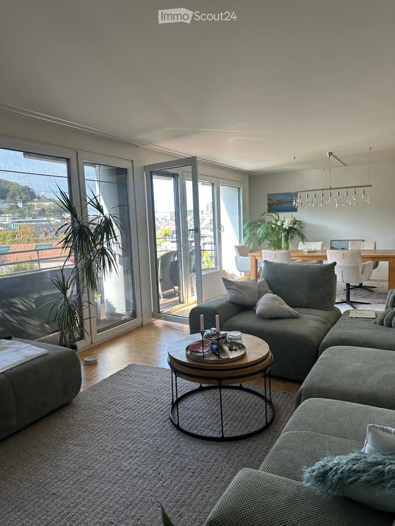 Modern living room with large windows and a view, featuring a grey sectional sofa, round coffee table, dining area with chairs and table, and potted plants.