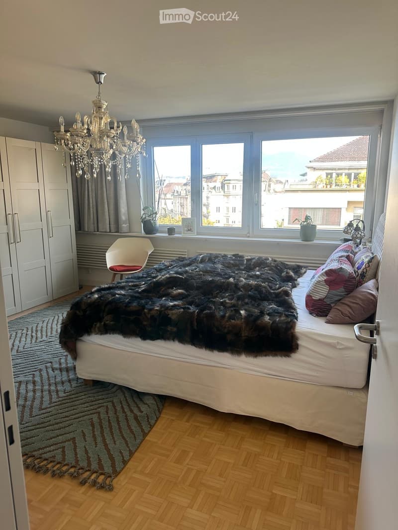 Cozy bedroom with wooden flooring, large windows, a bed with fur throw and colorful pillows, a white chair, a wardrobe, and a chandelier.