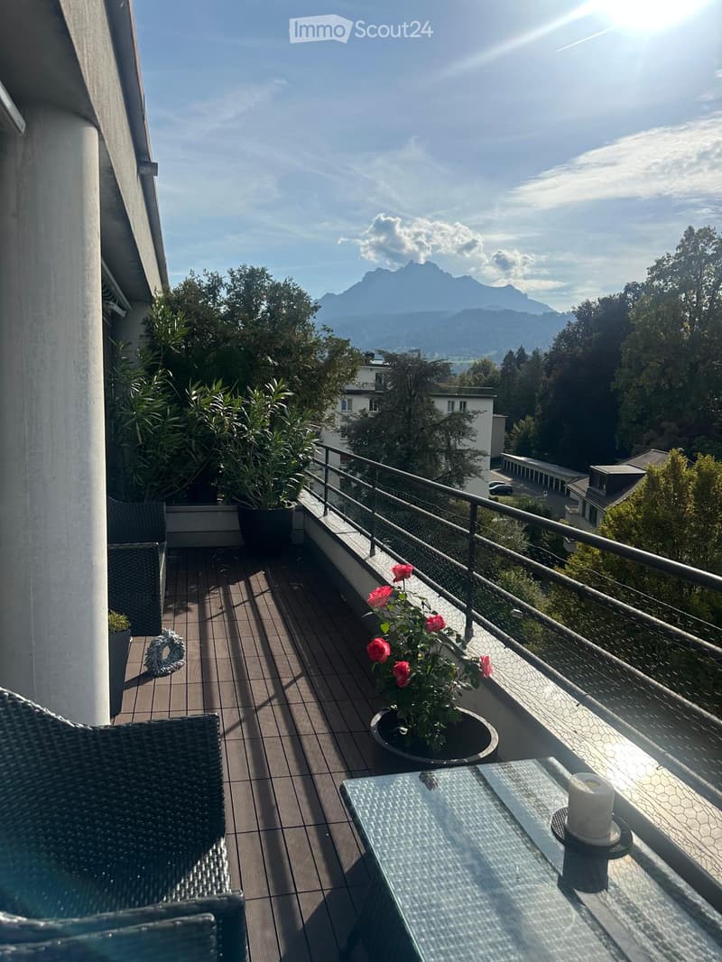 Balcony with wooden decking, glass table, wicker chairs, potted plants, and a scenic mountain view.