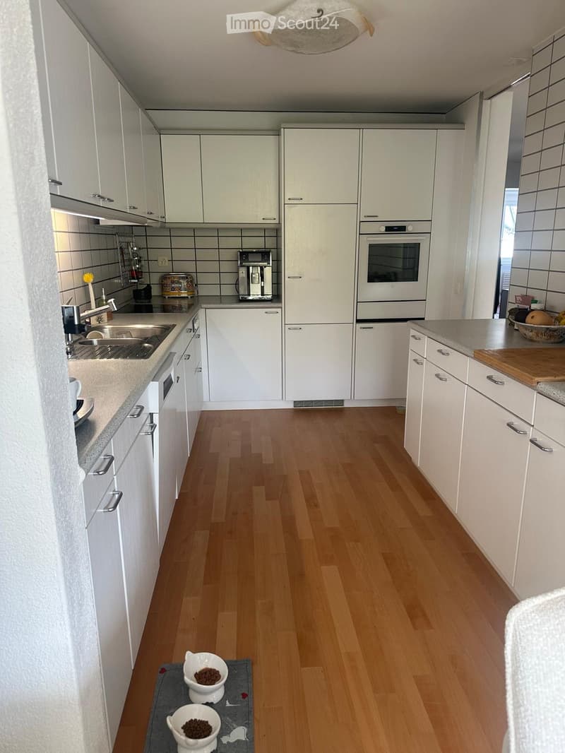 Modern kitchen with white cabinets, wooden flooring, stainless steel sink, oven, coffee maker, and toaster. Includes a pet feeding area with bowls on a mat.