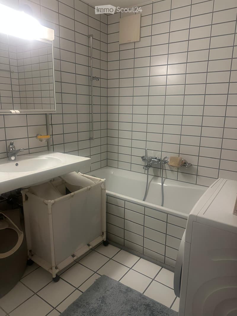 Bathroom with white tiled walls and floor, featuring a sink, bathtub, mirror, and washing machine. A small trash bin is placed under the sink, and a blue bath mat lies on the floor.