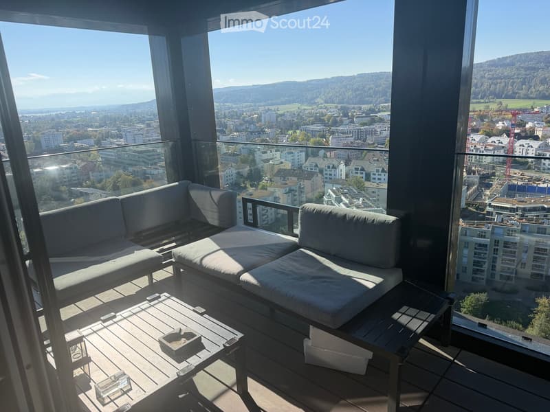 Modern balcony with large windows offering a panoramic city view, featuring a grey sectional sofa and a wooden coffee table.