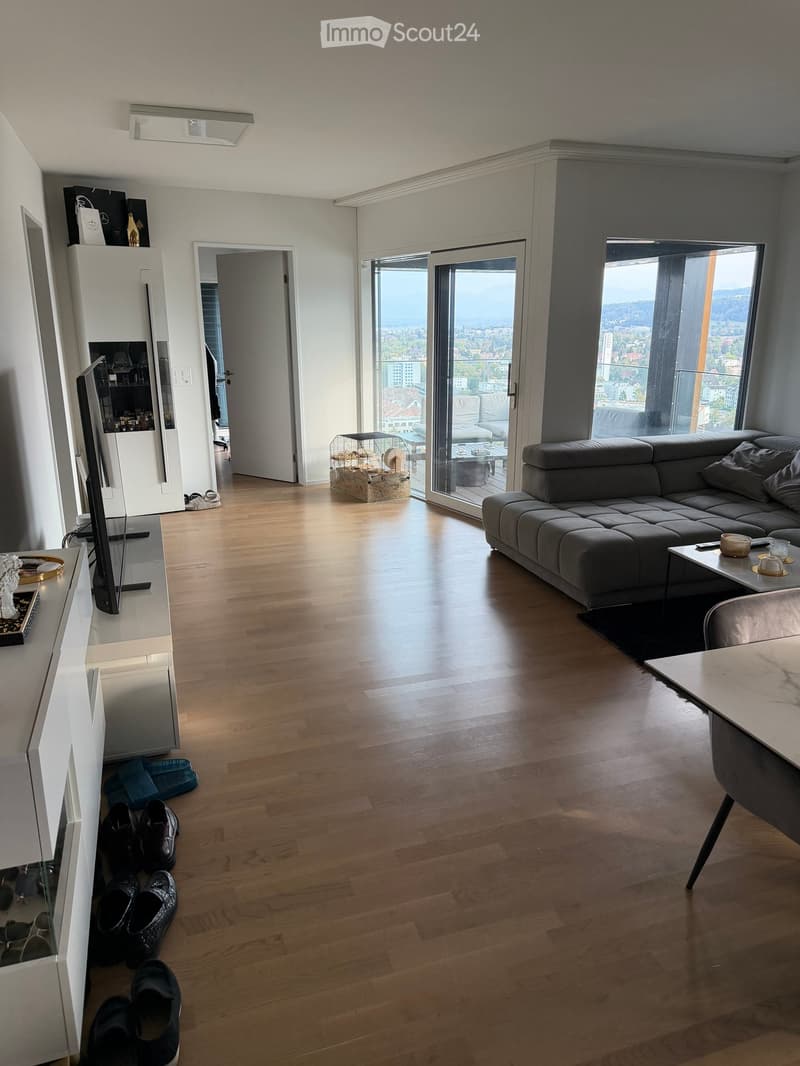 Modern living room with wooden flooring, large windows offering a city view, a grey sectional sofa, a coffee table, a TV stand, and a small table with chairs.