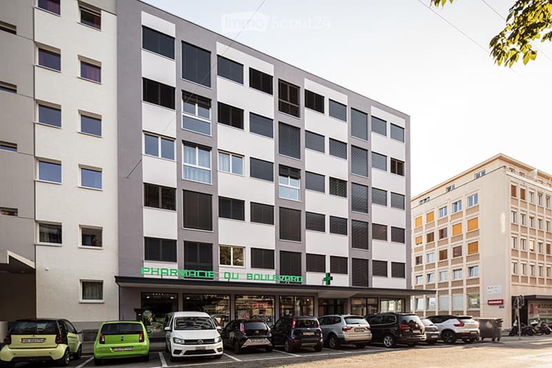 Multi-story building with modern facade, large windows, and a pharmacy sign reading "PHARMACIE DU BOULEVARD" at the entrance. Several cars are parked in front of the building.