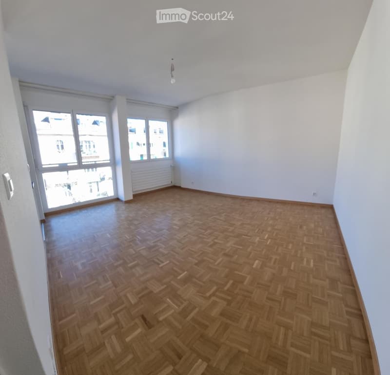 Empty room with wooden herringbone flooring, white walls, large windows, and a radiator. No furniture or equipment present.