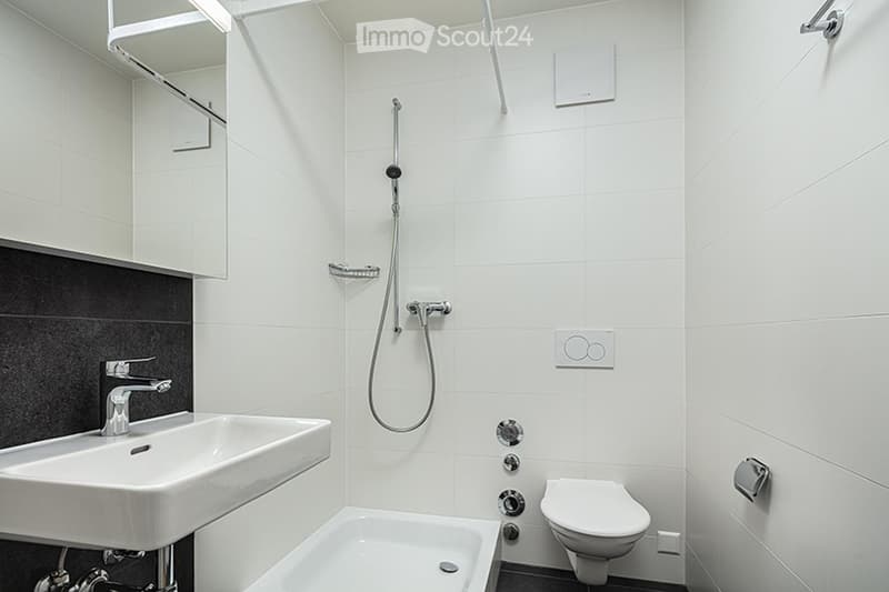 Modern bathroom with white tiles, a sink, bathtub, toilet, and a shower hose. Features a sleek design with a mirror and modern fixtures.