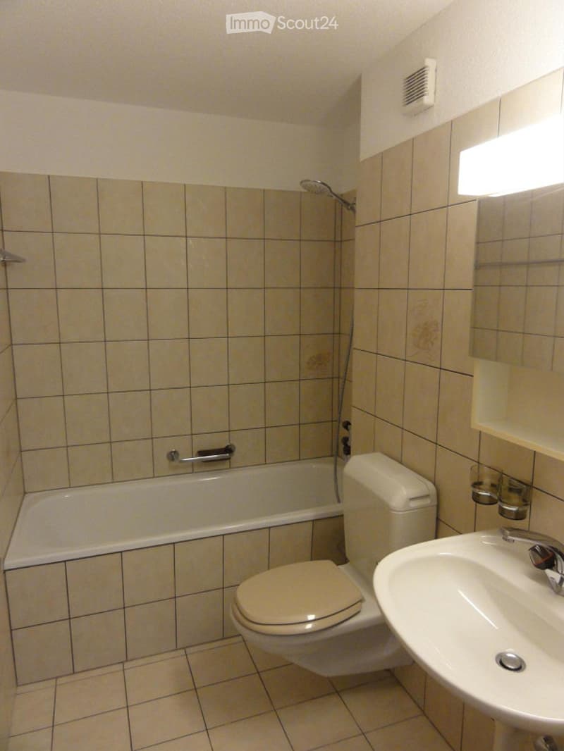 A small bathroom with beige tiled walls and floor, featuring a bathtub, toilet, sink, and a mirror above the sink.