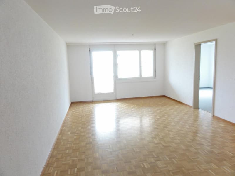 Empty room with wooden parquet flooring, white walls, large windows, and a sliding glass door leading outside.