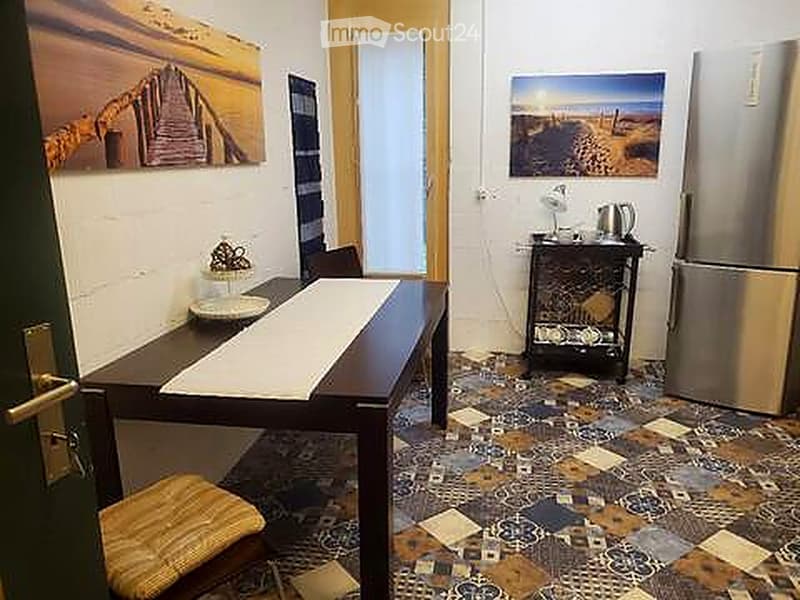 A small, furnished room with a dining table, chairs, a refrigerator, a small cabinet, and decorative picture frames on the walls.