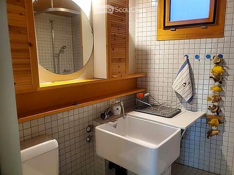 Cozy bathroom with a white sink, wooden vanity, mirror, and a small shower area. The walls are tiled with a grid pattern, and a towel hangs on the side.