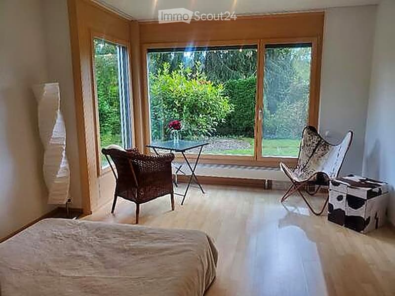 Bright, modern bedroom with large windows overlooking a garden, featuring a bed, wicker chair, small table, and decorative stool.