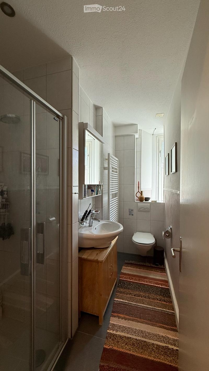 Modern bathroom with a sink, toilet, shower, and a small wooden cabinet. Features a striped rug, a mirror, and decorative items on the counter.