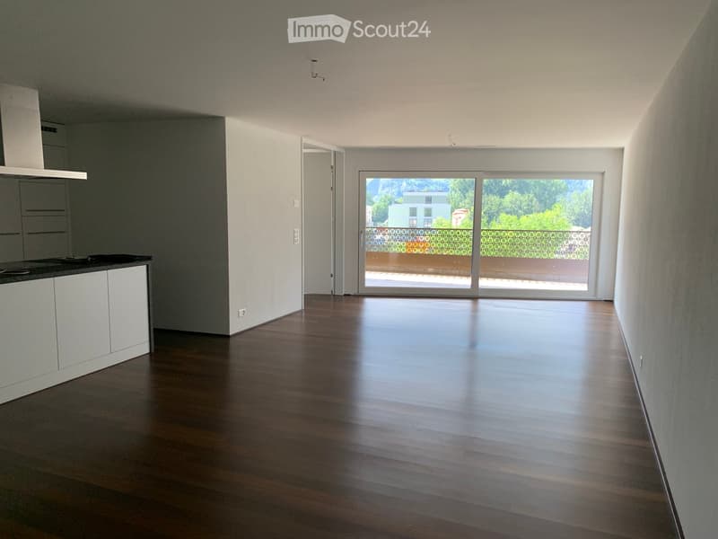 Empty room with wooden flooring, white walls, and a sliding glass door leading to a balcony with a view of greenery and a building.