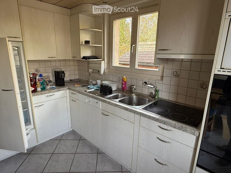 A modern kitchen with white cabinets, granite countertops, a stainless steel sink, and an electric stovetop. The kitchen features a refrigerator, coffee maker, and various cleaning supplies on the counter.