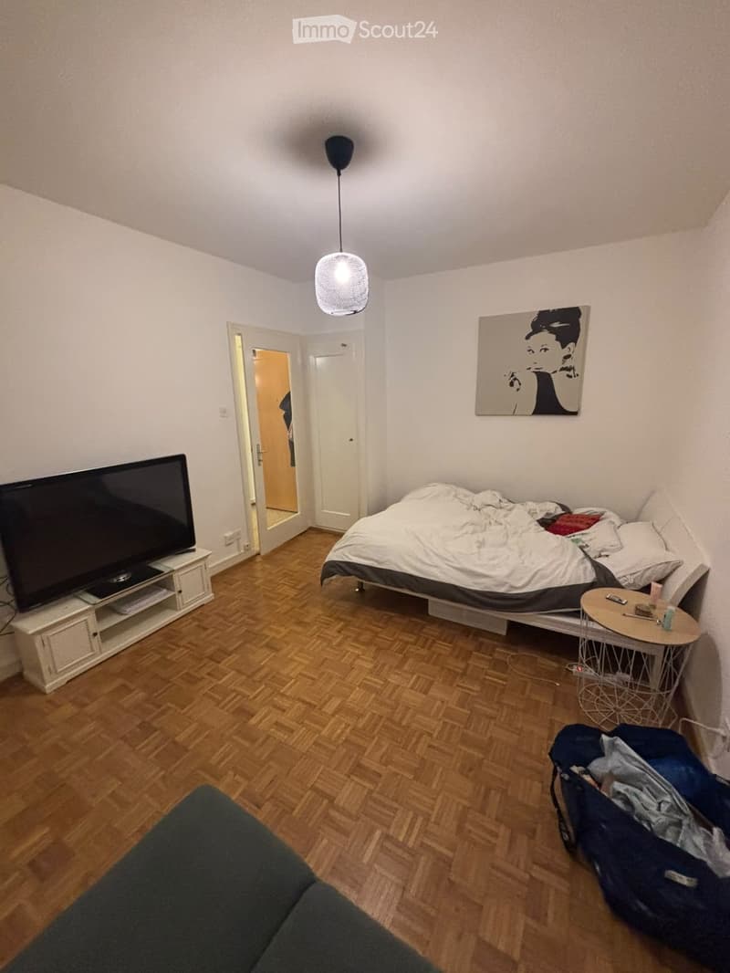 A modern bedroom with a wooden floor, a bed with white bedding, a nightstand, a small round table, a chair, a television on a stand, a hanging lamp, and a painting on the wall.