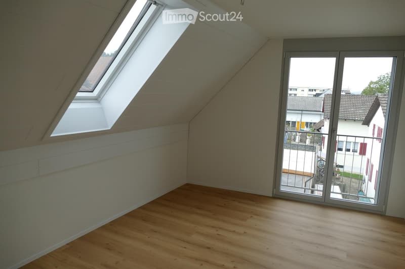 Empty attic room with wooden flooring, white walls, skylight windows, and a large sliding glass door leading to a balcony.