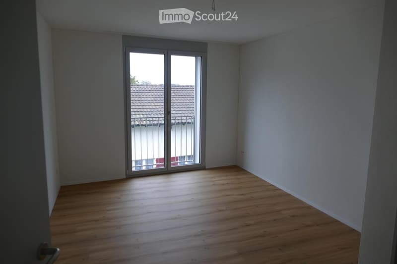 Empty room with wooden flooring and large sliding glass doors leading to a balcony. The walls are painted in neutral tones, and the space is well-lit with natural light.