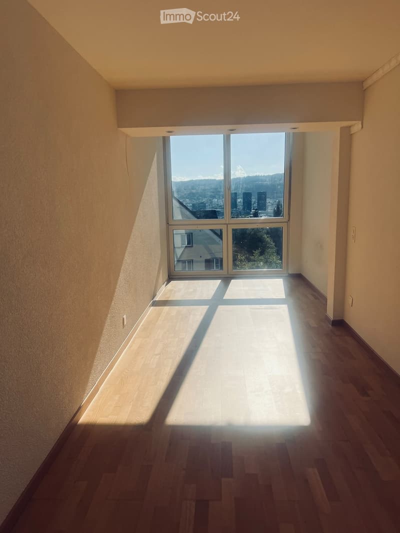 Empty hallway with wooden flooring, large window at the end, natural light streaming in, view of cityscape and mountains.