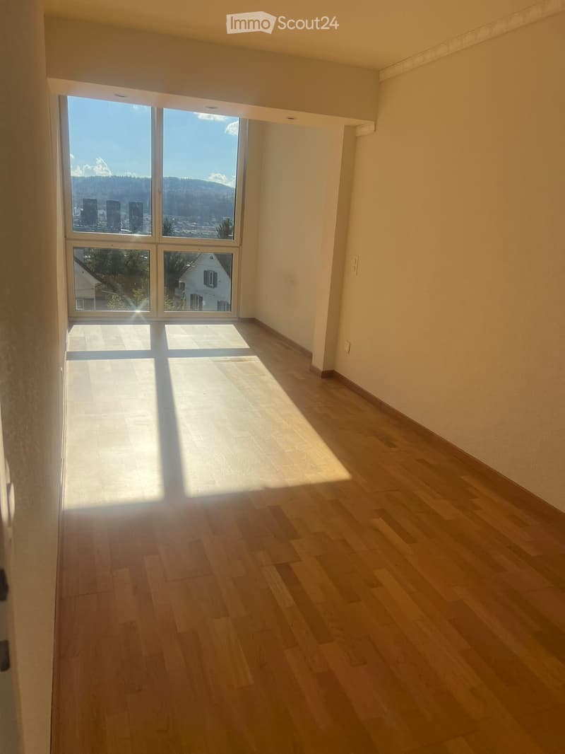 Empty room with wooden flooring and large windows offering a scenic view of mountains and cityscape.