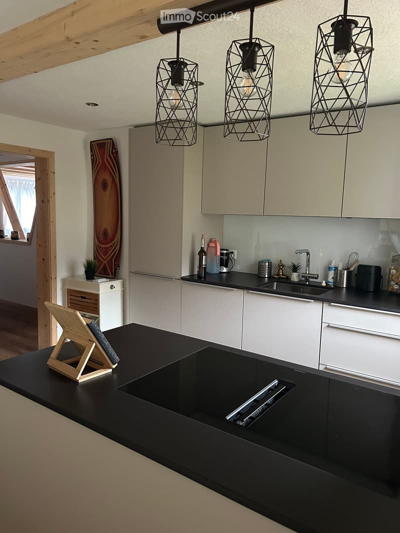 Modern kitchen with white cabinets, black countertops, and pendant lights. Features a central island with a sink, various kitchen items on the counters, and a small table with a chair in the corner.