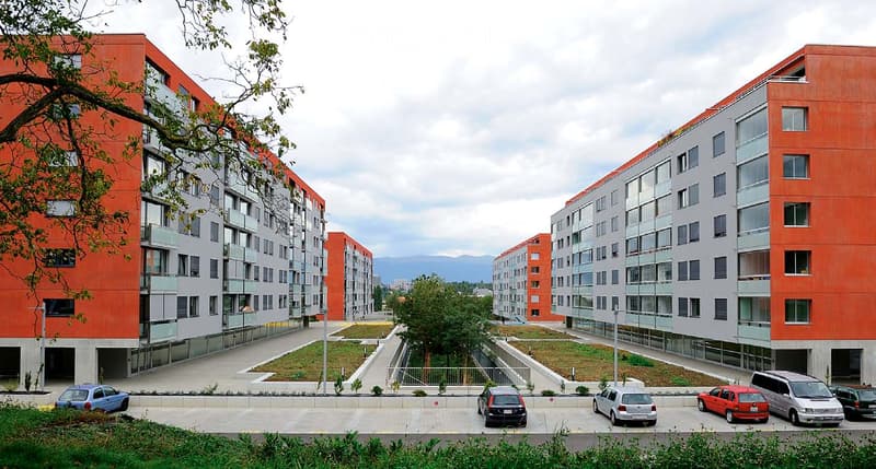 Modern apartment complex with red and grey facades, multiple floors, balconies, and a parking area with several cars.