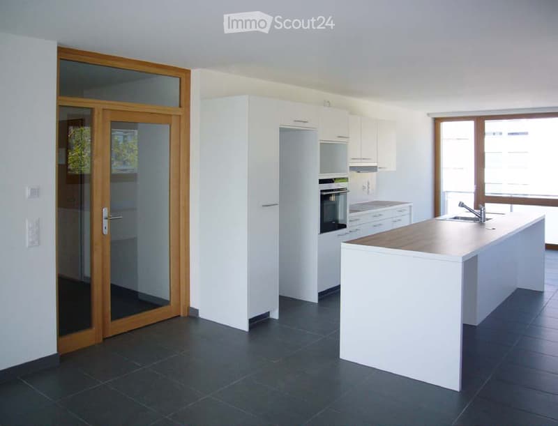 Modern kitchen with white cabinets, a central island, and a sink. Features include a large window, dark tiled flooring, and a sliding glass door leading outside.