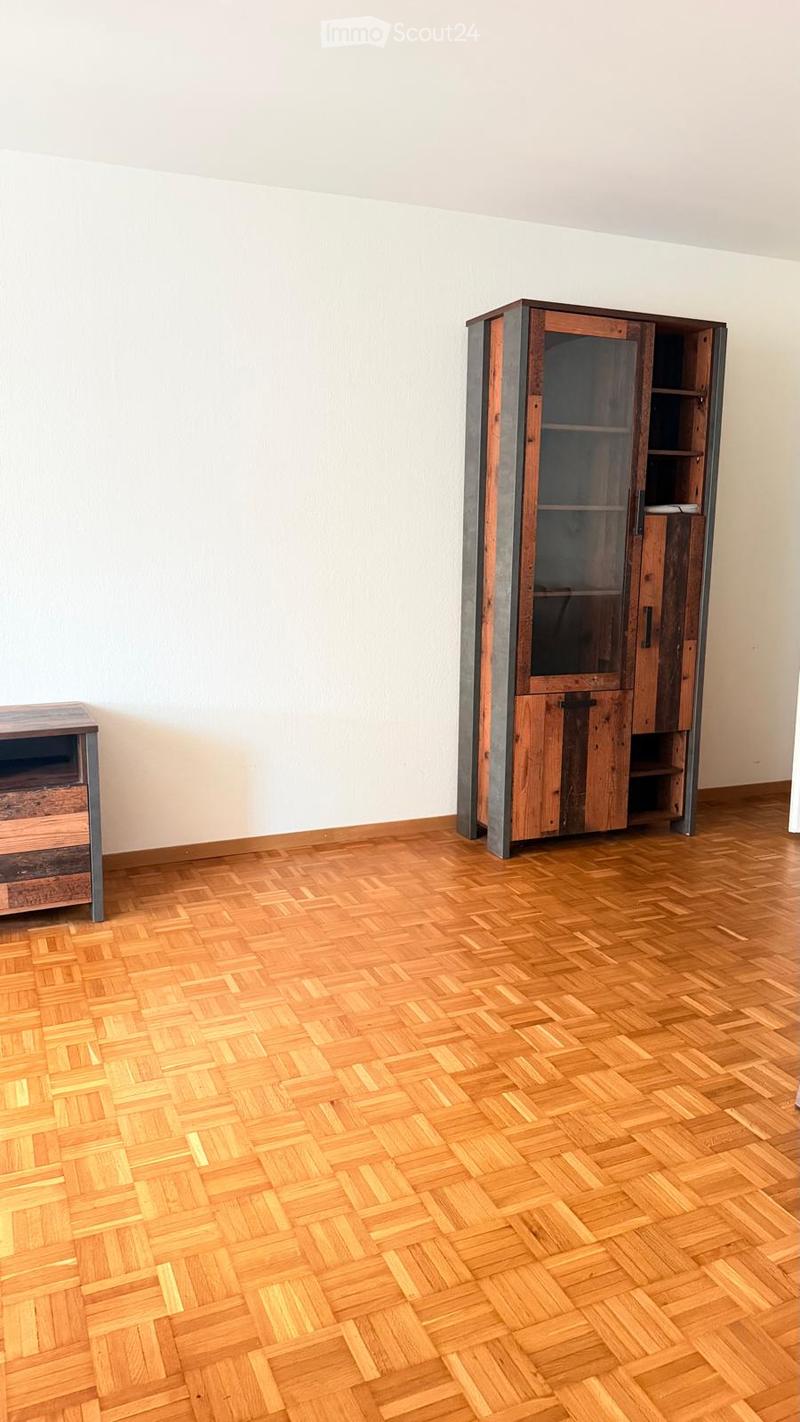A room with wooden herringbone flooring, white walls, and two pieces of furniture: a tall wooden cabinet with glass doors and a small wooden table.