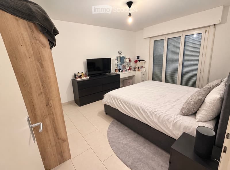 Modern bedroom with a large bed, nightstand, dresser, mirror, and television. The room features large windows with blinds and a sleek, contemporary design.