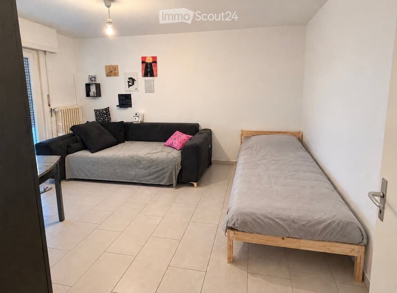 A small, well-lit room with a sofa, a single bed, a small table, shelves, and picture frames on the wall.
