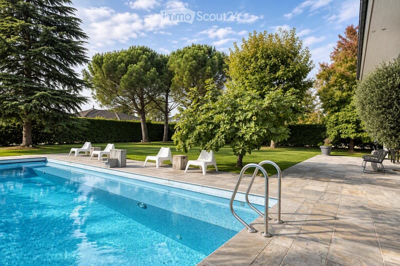 A well-maintained garden with a swimming pool, surrounded by lush greenery, trees, and neatly trimmed hedges. The pool area features lounge chairs and a small table, with a paved patio extending to the right side.