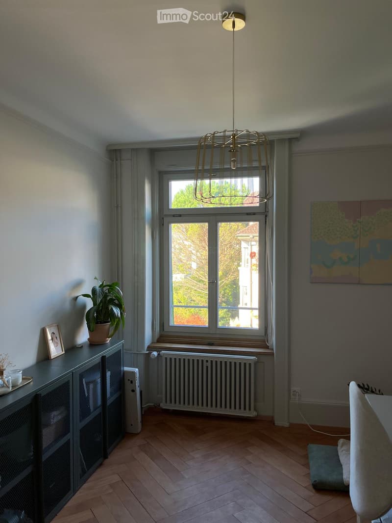 A modern living room with wooden flooring, large windows, a hanging lamp, a cabinet with a picture frame and potted plant, and a chair near the window.