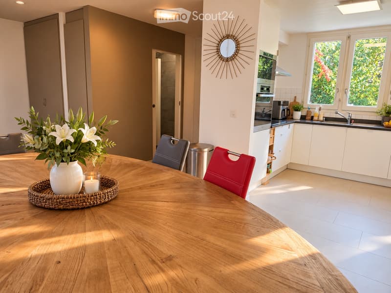 Bright, modern dining area with a wooden dining table, red and grey chairs, a vase with white lilies, and a candle. Adjacent kitchen features white cabinets, a sink, oven, coffee maker, and a decorative mirror on the wall.