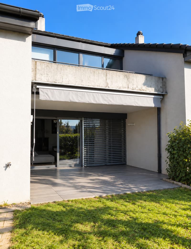 Modern two-story house with large windows, a balcony, and a covered entrance area. The house features a clean, contemporary design with a raised ground floor and a well-maintained lawn in the foreground.