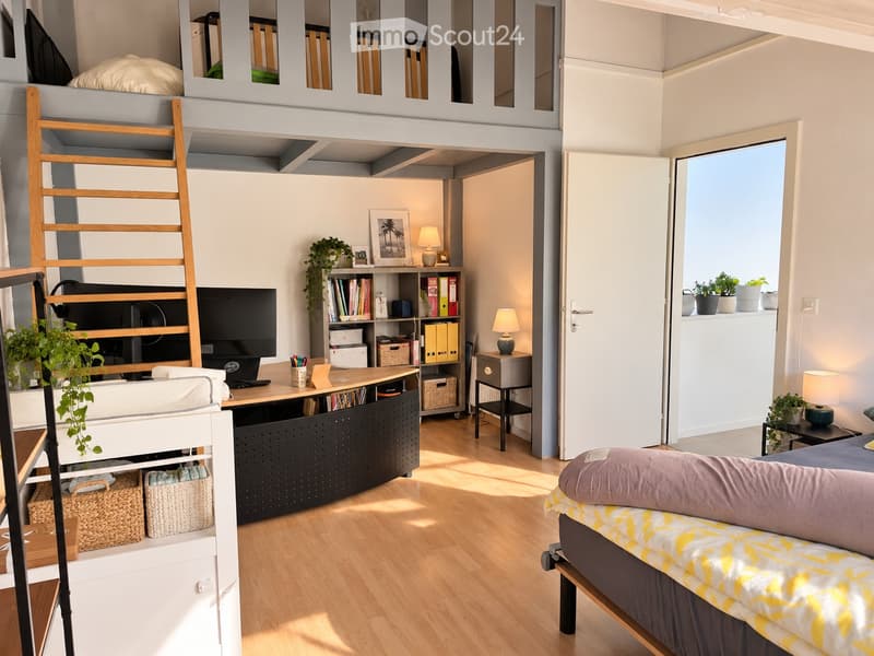 A modern, multi-functional room with a raised bed area, a desk with a laptop and screen, a sofa, and a bookshelf. The room features wooden flooring, large windows with a view, and several potted plants.