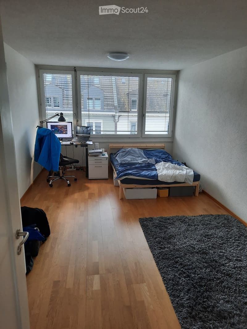 A small, well-lit bedroom with a wooden floor, a single bed with blue bedding, a desk with a laptop and chair, a storage unit, and a large shaggy rug near the bed.