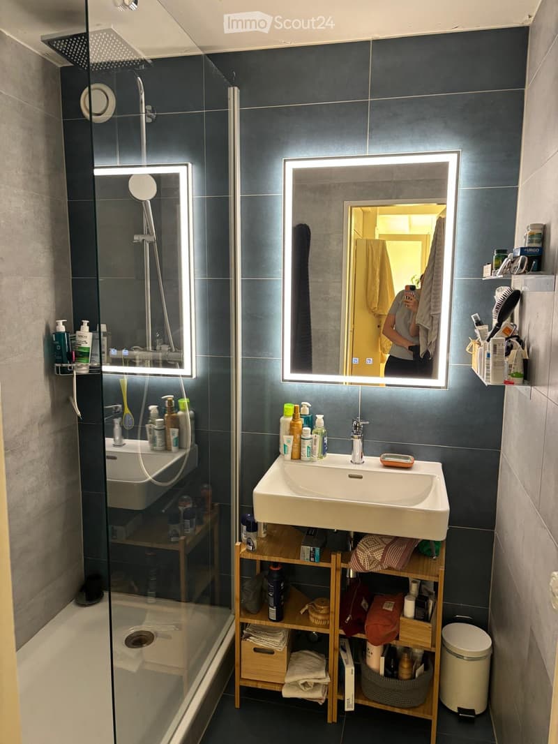 Modern bathroom with dark tiled walls, a large illuminated mirror, a sink with multiple toiletries, a wooden shelf holding towels and items, and a glass-enclosed shower area.