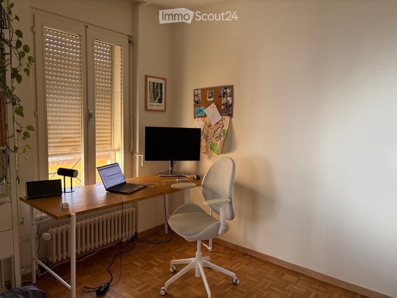 A modern, well-lit home office with a wooden desk, laptop, ergonomic chair, and large window with blinds.