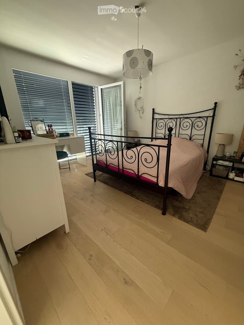 Bright bedroom with wooden flooring, large windows with blinds, a metal-framed bed with pink bedding, a nightstand, a small table, and a hanging lamp.