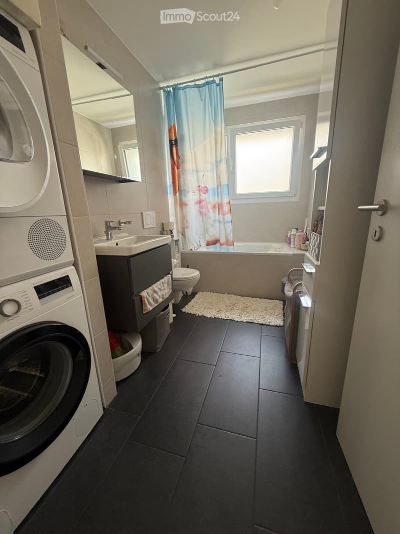 Modern bathroom with dark tiled floor, white walls, a sink with a faucet, a toilet, a bathtub with a shower curtain, a washing machine, a mirror, and a small white rug in front of the bathtub.