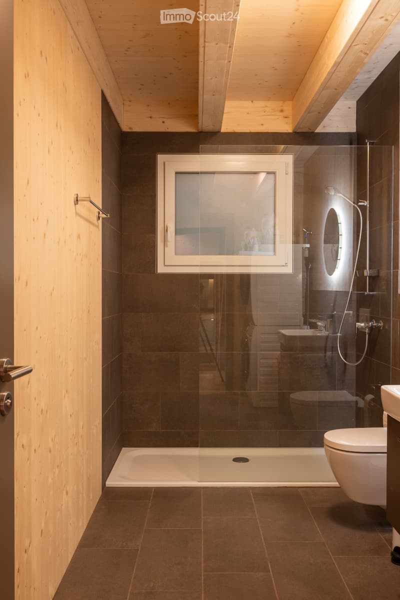 Modern bathroom with dark tiled walls and floor, a large bathtub, a toilet, a glass-enclosed shower area, and a window above the bathtub.