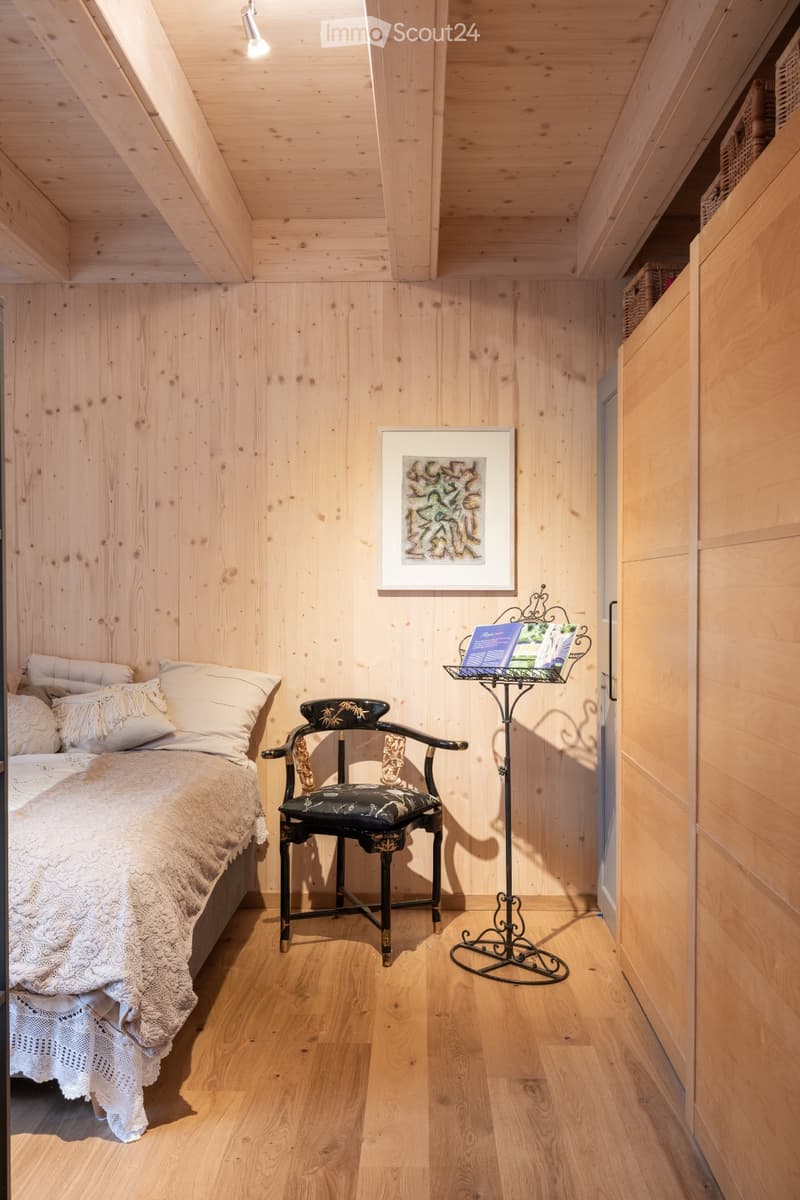Cozy attic bedroom with wooden walls and ceiling, featuring a bed with white bedding, a decorative chair, a small table with a magazine rack, and a framed artwork on the wall.