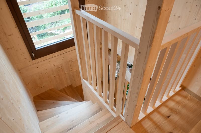 Wooden staircase with light-colored wooden railings and steps, leading up to a landing area with a window overlooking greenery.