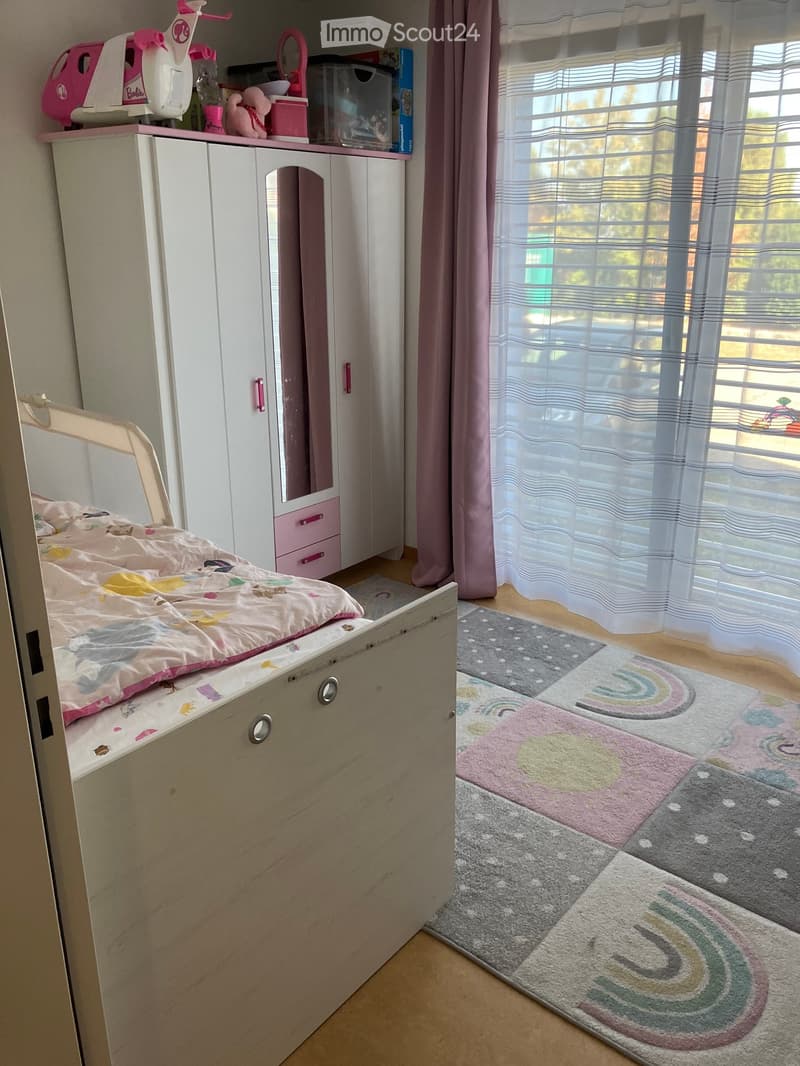 A child’s bedroom with a bed, colorful bedding, a wardrobe with a mirror, a toy on top, and a rainbow-patterned rug.