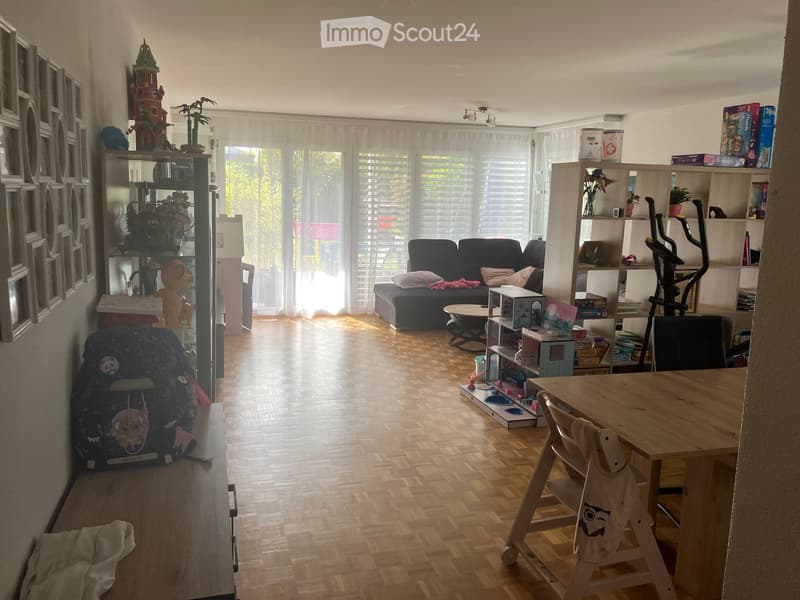Bright living room with wooden flooring, large windows with white curtains, a black sofa with pillows, a small round table, a wooden dining table with chairs, shelves with books and toys, and an exercise bike in the corner.