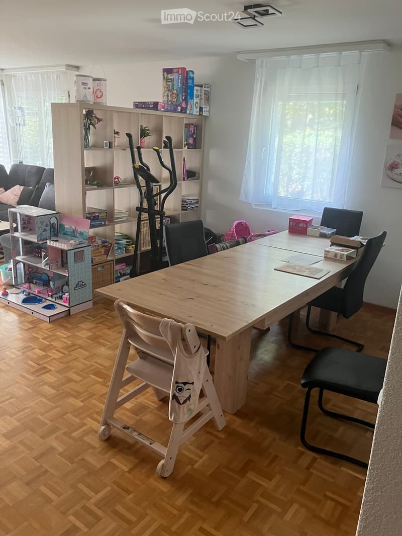 A well-lit room with wooden flooring, a large wooden table with chairs, a sofa, a bookshelf filled with books and toys, an exercise bike, and a toy house on the floor.