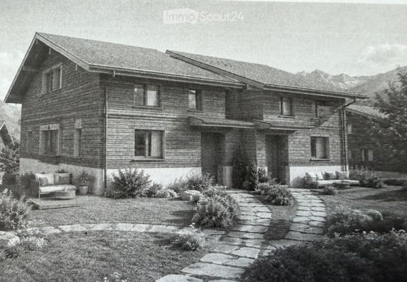 Two-story brick house with a sloped roof, multiple windows, and a stone pathway leading to the entrance. The house is surrounded by a well-maintained garden with shrubs and small seating areas.
