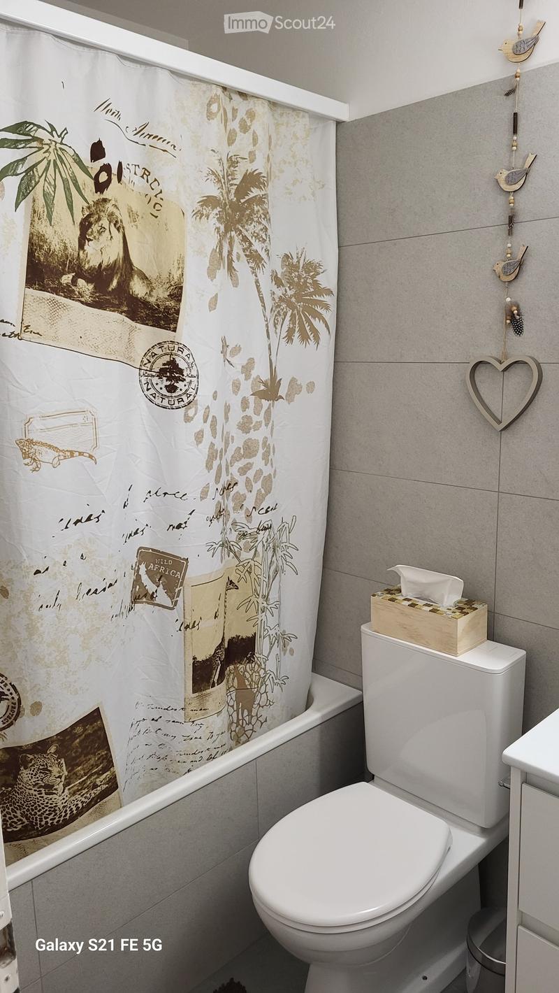 Modern bathroom with a white toilet, bathtub, and a decorative shower curtain featuring a safari theme. A tissue box is placed on top of the toilet tank, and a heart-shaped wall decoration is visible.