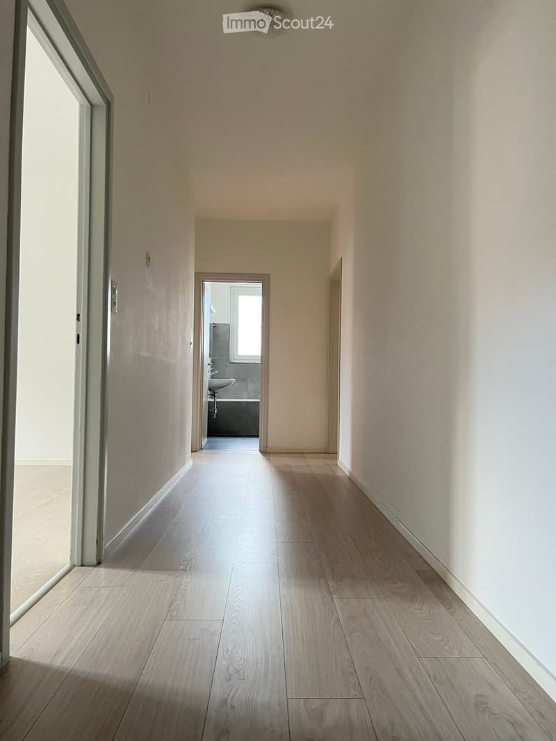 Empty hallway with light wooden flooring and white walls, featuring a glass door on the left and a partially visible bathroom at the end.
