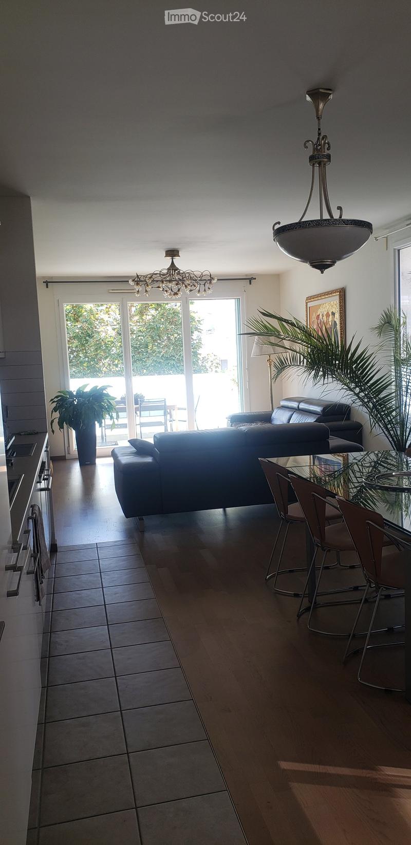 Open-plan living and dining area with a dark leather sofa, glass dining table with chairs, chandelier, floor lamp, and potted plants near large windows leading to a balcony.