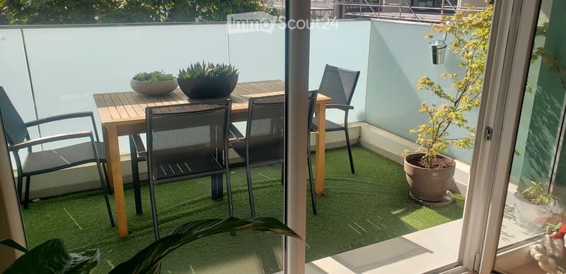 Balcony with artificial grass, wooden table, black chairs, and several potted plants.